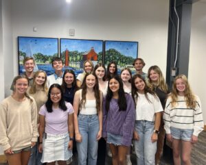 The 19 member 2024-2025 Youth Advisory Council stands in a group. Nearly all the high school-age students are smiling, and they are standing in front of a painting of the Grand Haven Pier.